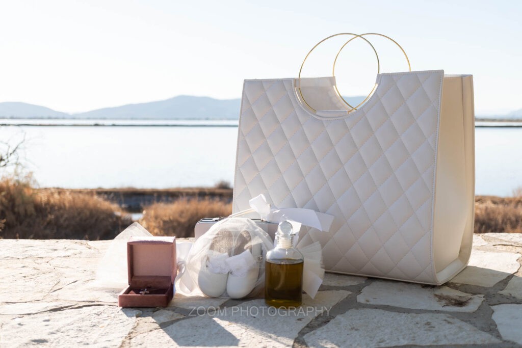 Λαδικό, παπουτσάκια, σταυρός και λευκή τσάντα βάπτισης σε πέτρινο τοίχο, με θέα τη λιμνοθάλασσα