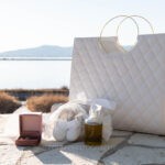 Λαδικό, παπουτσάκια, σταυρός και λευκή τσάντα βάπτισης σε πέτρινο τοίχο, με θέα τη λιμνοθάλασσα
