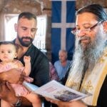 Η μικρή χαμογελά στην αγκαλιά του νονού της, δίπλα στον ιερέα, μέσα στην Παναγία Φοινικιά