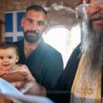 Ο νονός κρατάει τη μικρή μπροστά στον ιερέα, μέσα στο πέτρινο εκκλησάκι