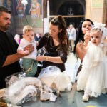 Ο Χρήστος κρατά τον γιο του και η Φωτεινή στέκεται όρθια με το λευκό δαντελένιο φόρεμα την ώρα που τα ντύνουν