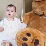 Η Φωτεινή με το λευκό βαπτιστικό φόρεμα και τον σταυρό της κάθεται δίπλα σε μεγάλο λούτρινο αρκουδάκι