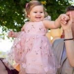 Η Μαριάνθη χαμογελά με ροζ φορεματάκι με κεντημένα λουλούδια, ψηλά στην αγκαλιά των δικών της