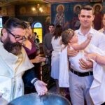 Ο νονός κρατά τη Μαριάνθη τυλιγμένη στο λευκό πανί μετά την κολυμβήθρα