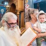 Ο Γεράσιμος με το λευκό καπέλο στην αγκαλιά της νονάς, δίπλα στον ιερέα